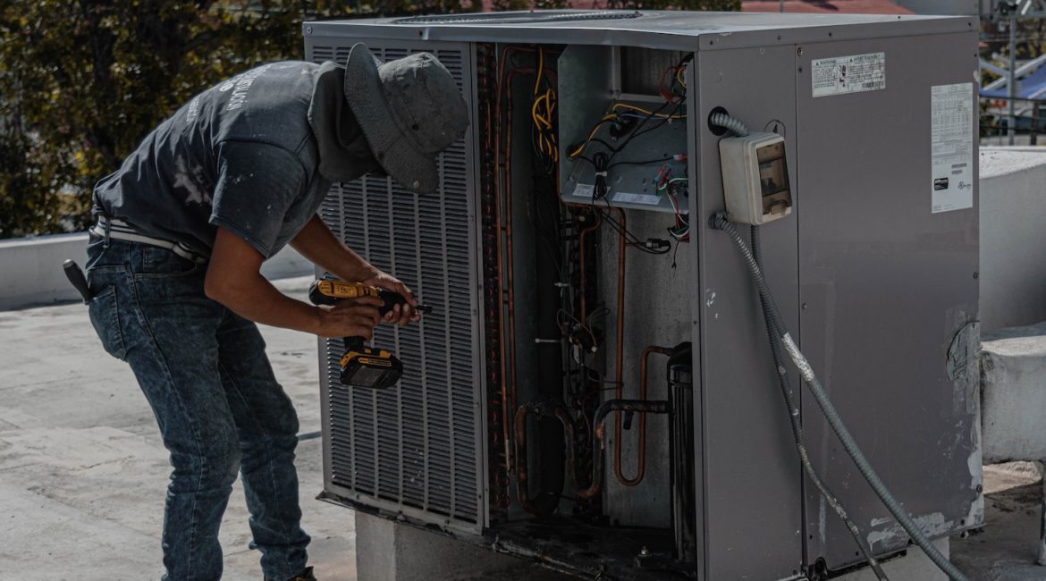 A worker in a hat using a drill to repair an air conditioning unit on a rooftop under sunligh