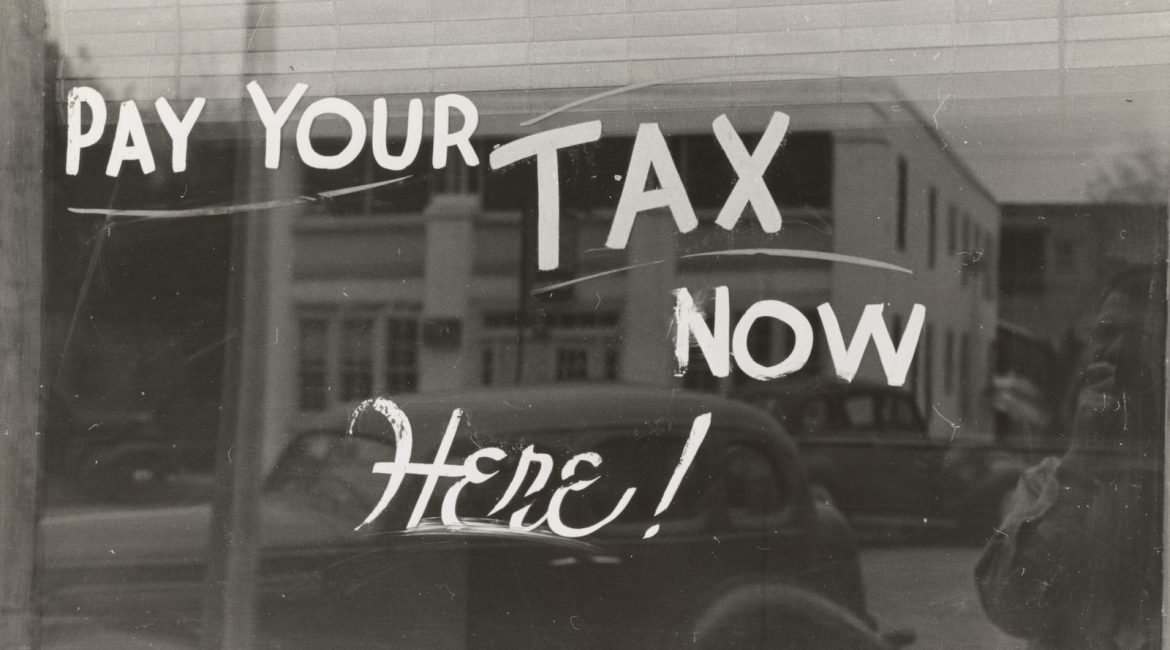 Black-and-white photo of a glass window with a hand-painted sign reading “Pay Your Tax Now Here!” reflecting a vintage car