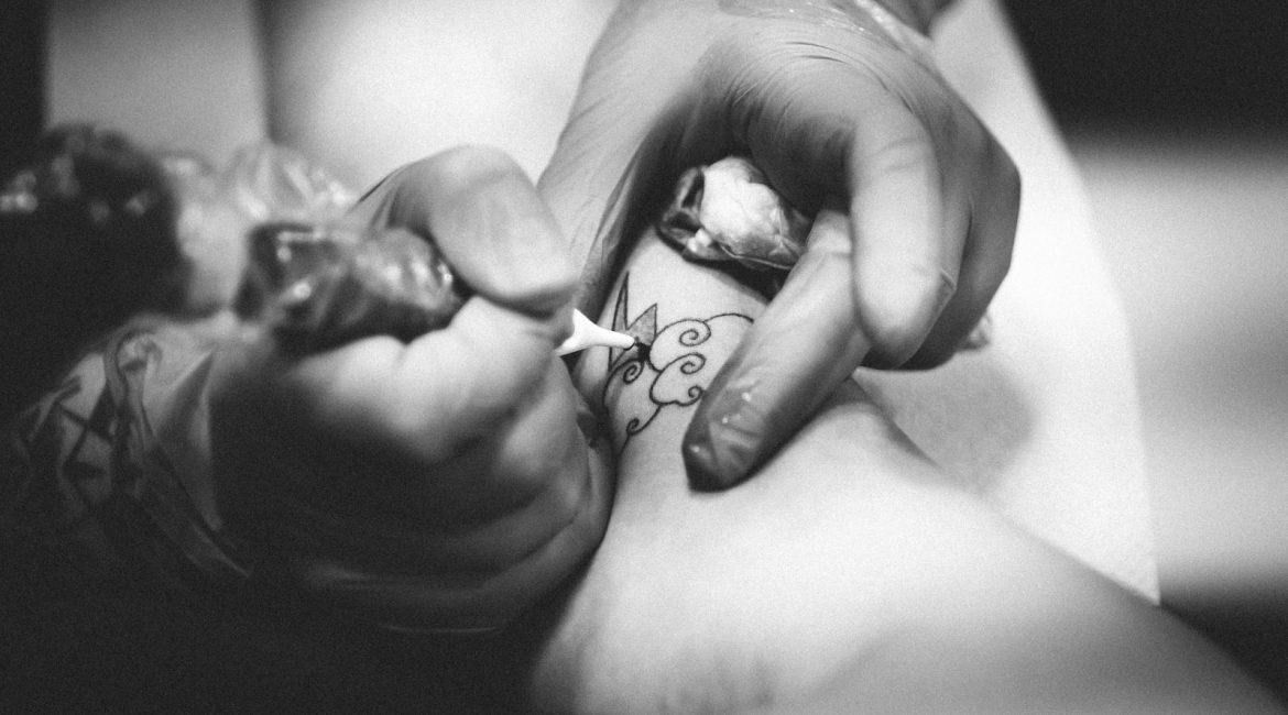 Black-and-white close-up of a tattoo artist working on a design on a person's leg with a tattoo machine