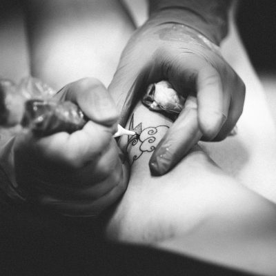 Black-and-white close-up of a tattoo artist working on a design on a person's leg with a tattoo machine