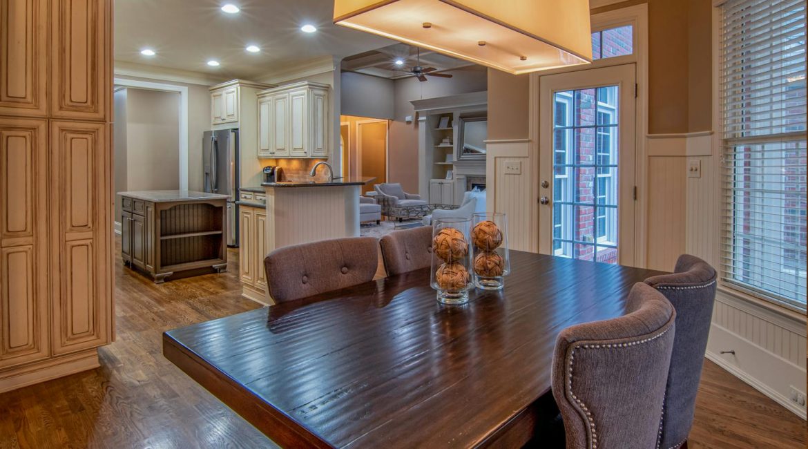 Bright and spacious dining area with a wooden table, grey upholstered chairs, and decorative glass jars, leading into a modern kitchen with ample lighting and a sliding door to the outside