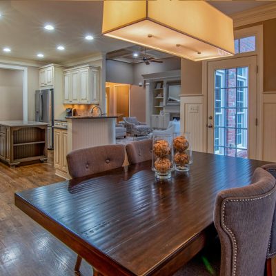 Bright and spacious dining area with a wooden table, grey upholstered chairs, and decorative glass jars, leading into a modern kitchen with ample lighting and a sliding door to the outside