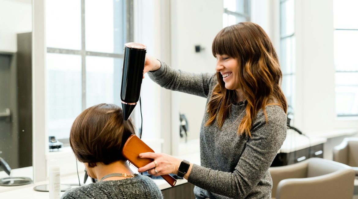 Hairstylist blow-drying a client's hair in a bright salon, both smiling during the process