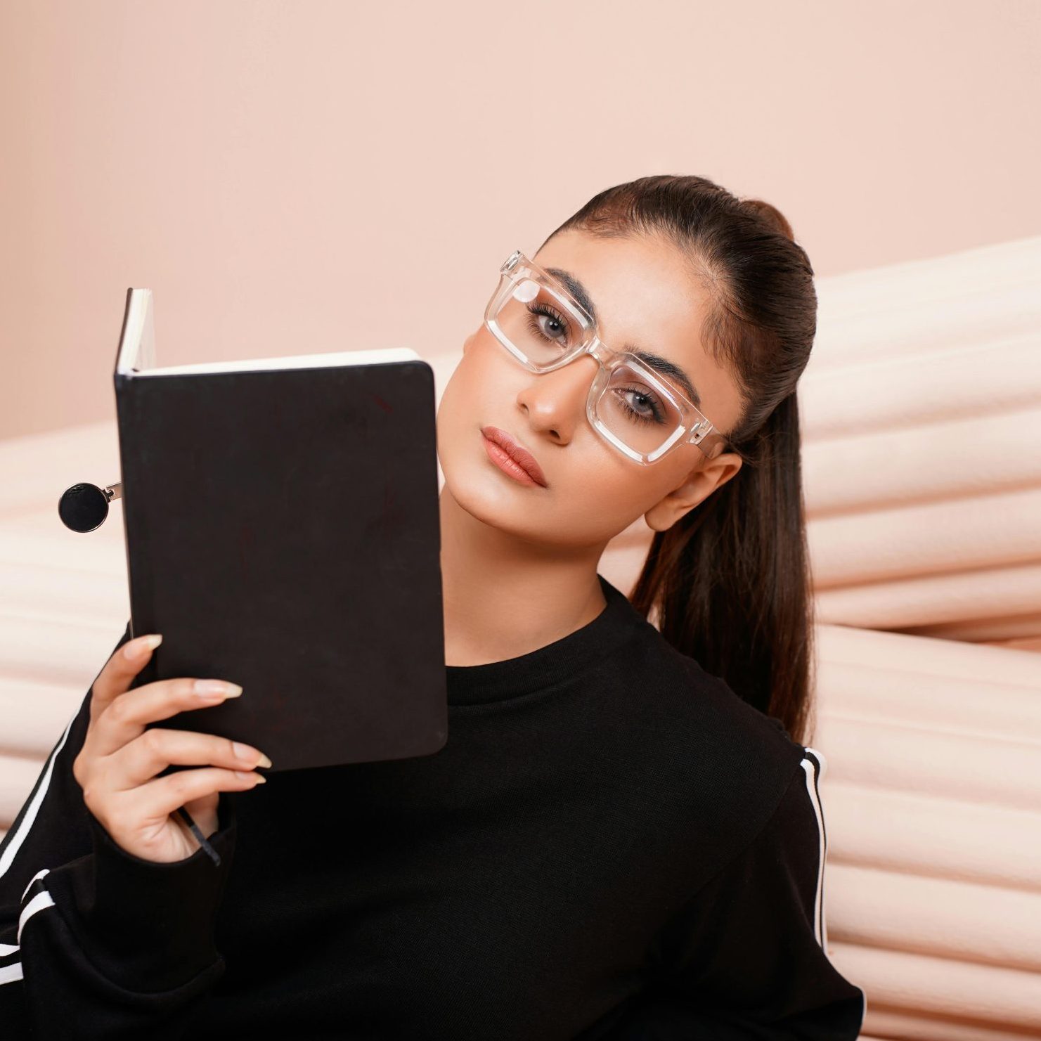 A young woman wearing clear glasses and a black sweatshirt, holding a closed notebook while looking thoughtfully at the camera, against a soft, neutral background