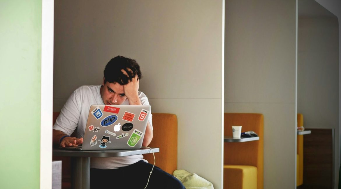 A person with curly hair, wearing a white t-shirt, sitting at a table and looking frustrated while working on a laptop covered with stickers, in a modern, minimalist setting