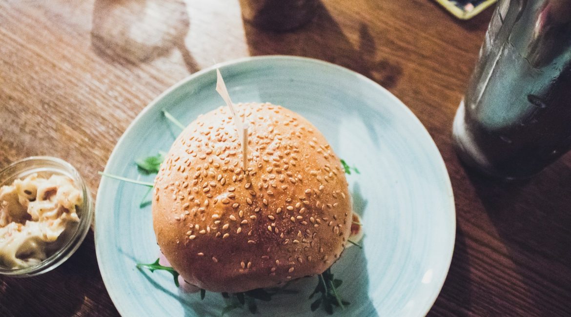 A sesame seed bun sandwich with fillings, placed on a blue plate with a side of creamy dip, set on a wooden table with a glass of drink in the background